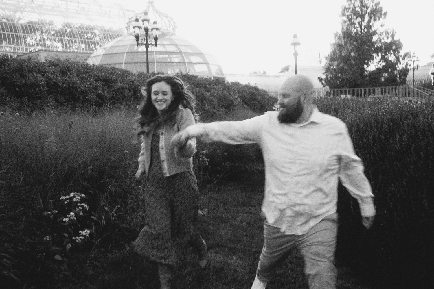 Black and white photo of a couple holding hands and dancing outdoors near a greenhouse garden.