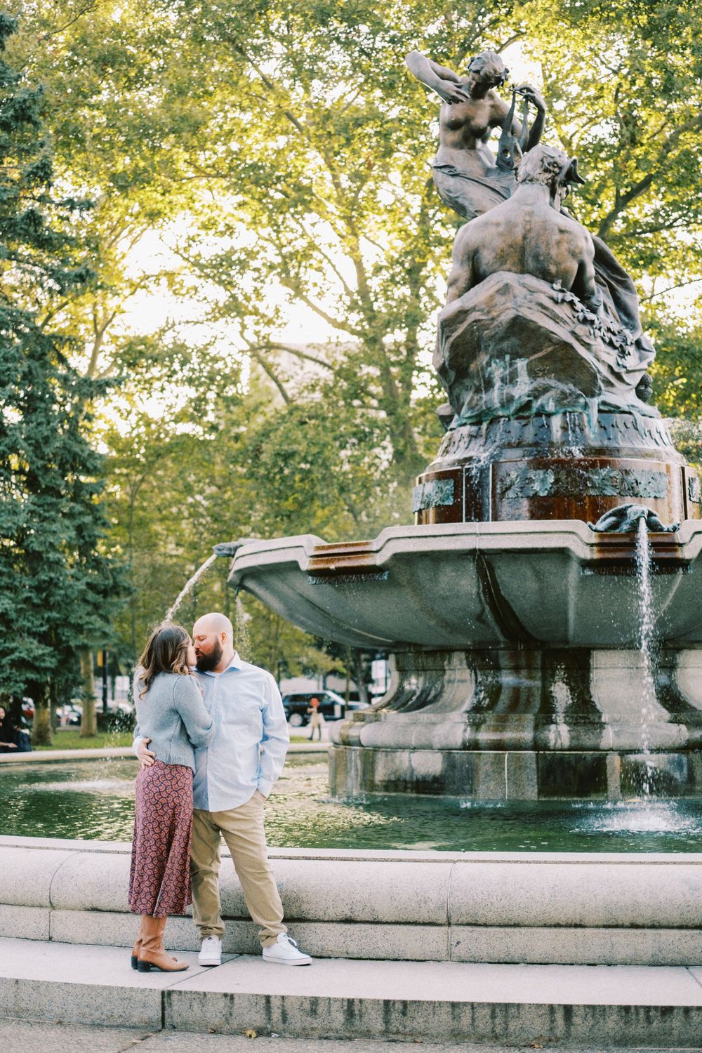 Couple embracing romantically beside an ornate fountain statue in a lush green park on a sunny day.