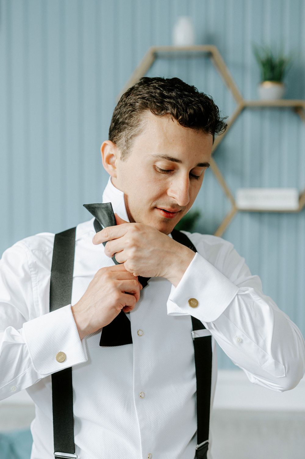 A groom in a white dress shirt with suspenders adjusts his black bow tie while getting ready for his wedding.
