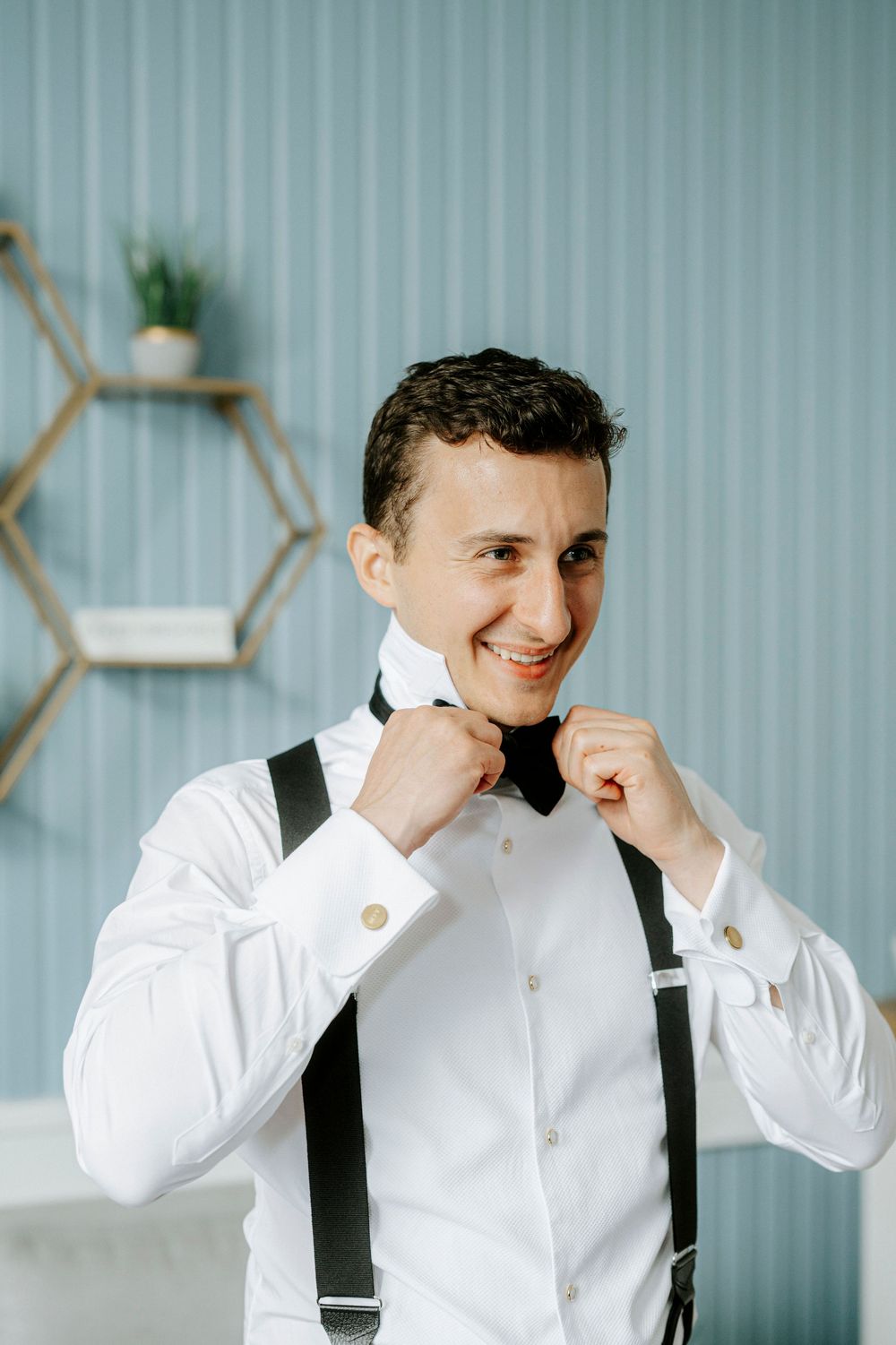 Smiling groom in white shirt adjusting black bow tie with suspenders, getting ready for wedding day.