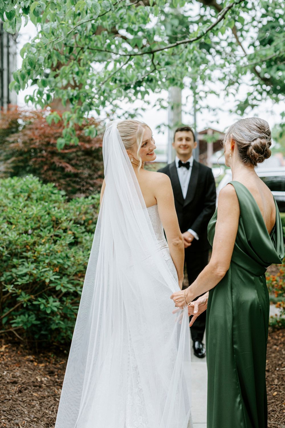 Bride in white gown and veil holds hands with bridesmaid in green dress, groom in tuxedo smiling in background.