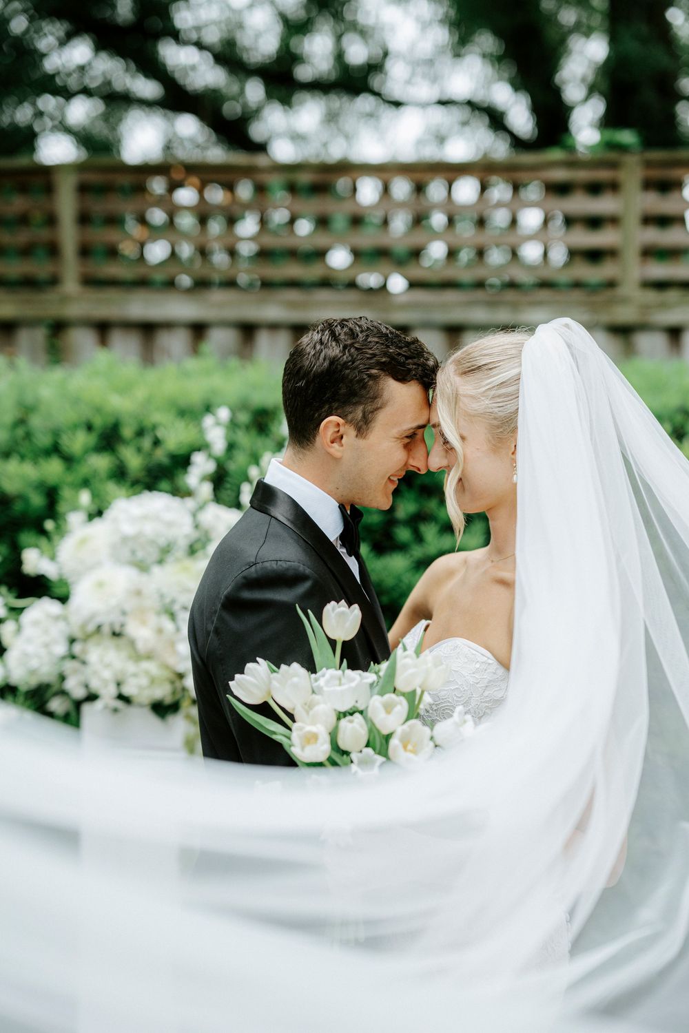 Bride and groom touching foreheads, bride holding white tulip bouquet with veil flowing, lush garden backdrop.