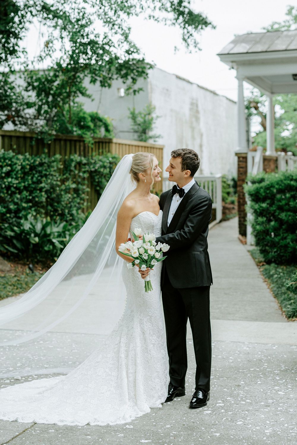 Bride in strapless lace gown with cathedral veil smiles at groom in black tuxedo on garden pathway.