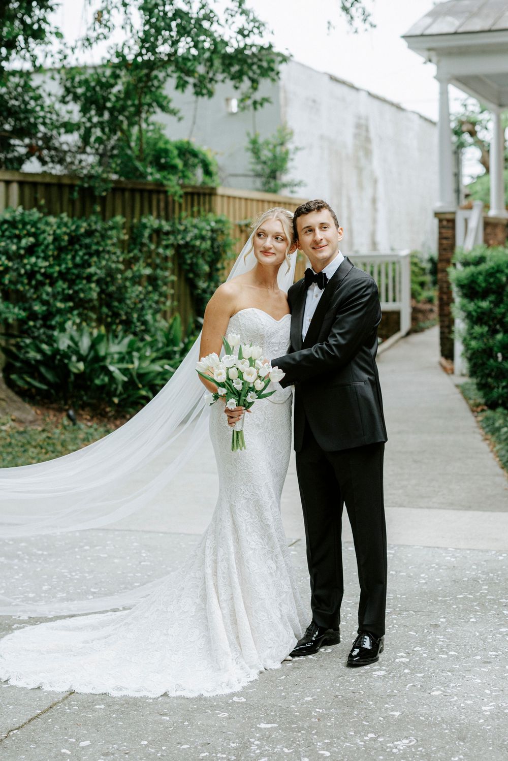 Bride in strapless lace gown with long veil holds white bouquet beside groom in black tuxedo on outdoor pathway.