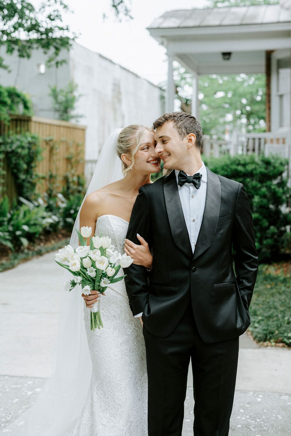 Bride in lace gown holding white tulip bouquet smiles with groom in black tuxedo outdoors.