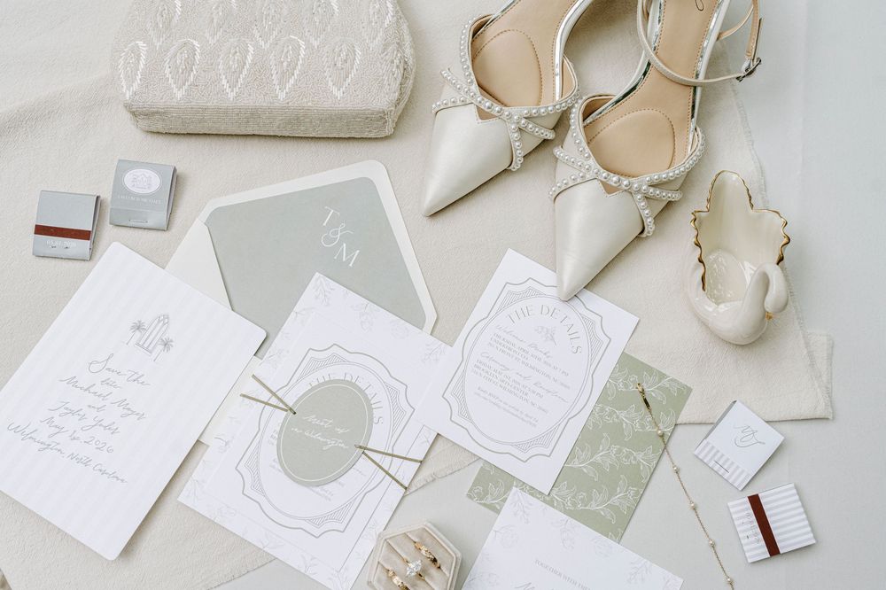 Flat lay of wedding stationery suite, ivory heels, clutch purse, and sage green invitation details on neutral background.
