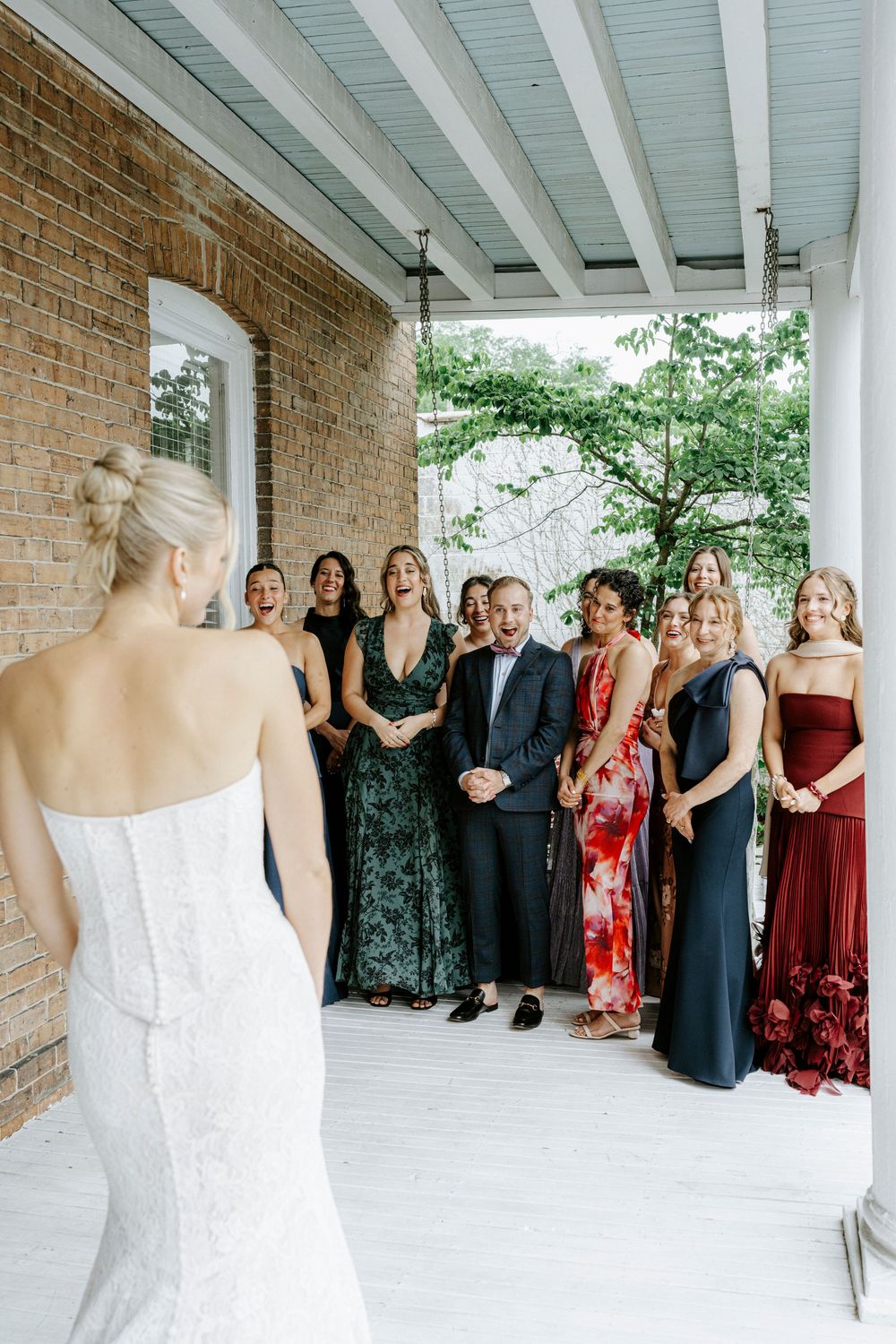 Bride reveals her wedding dress to excited bridesmaids and groomsman on a white porch during first look.