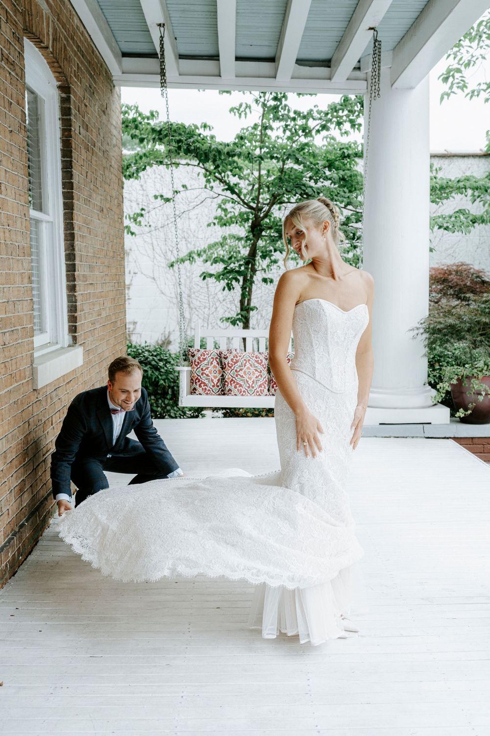 Groom adjusting bride's white mermaid wedding dress train on a classic Southern porch.