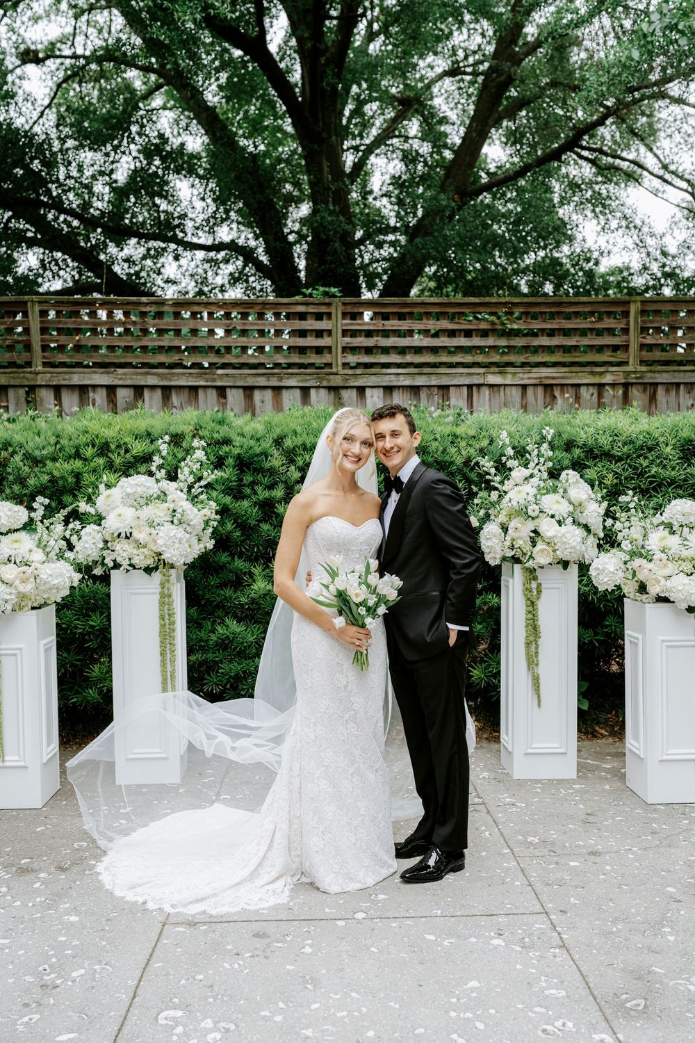Bride and groom posing together outdoors at wedding ceremony with white floral arrangements on pedestals and lush greenery.