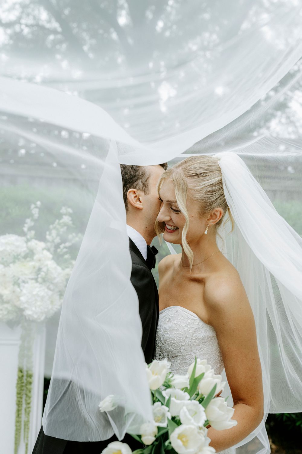 Bride and groom laughing under flowing white veil, bride holding white tulip bouquet at outdoor wedding.