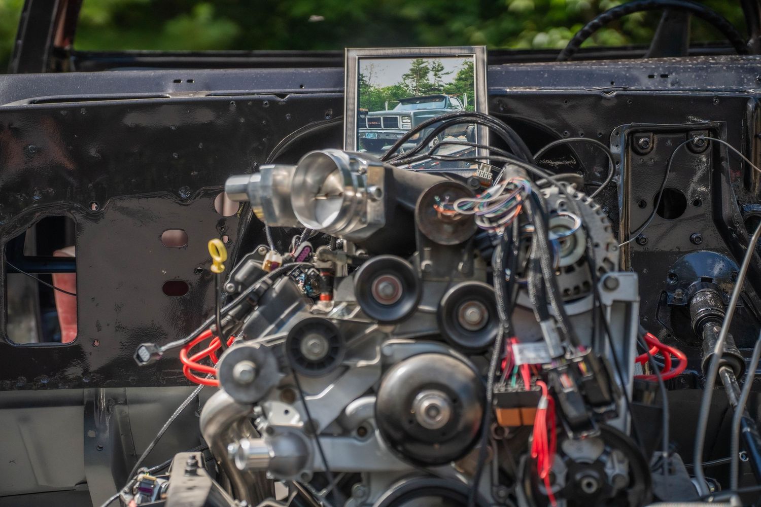 Exposed car engine with wiring and belts visible, with a small framed photo mounted above it outdoors.