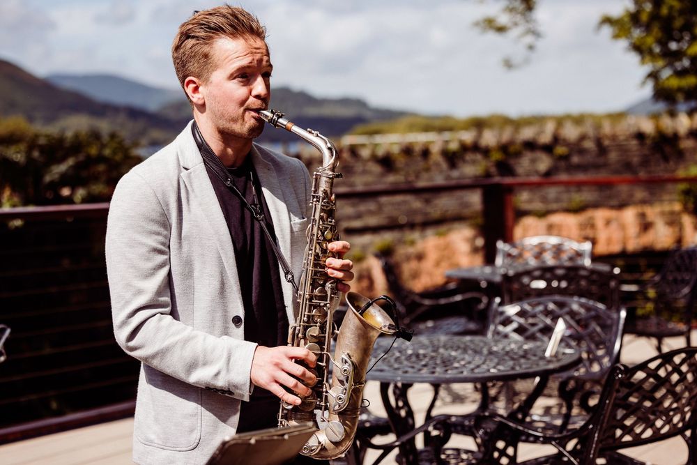 Young man in gray blazer playing saxophone outdoors on sunny terrace with mountain views.