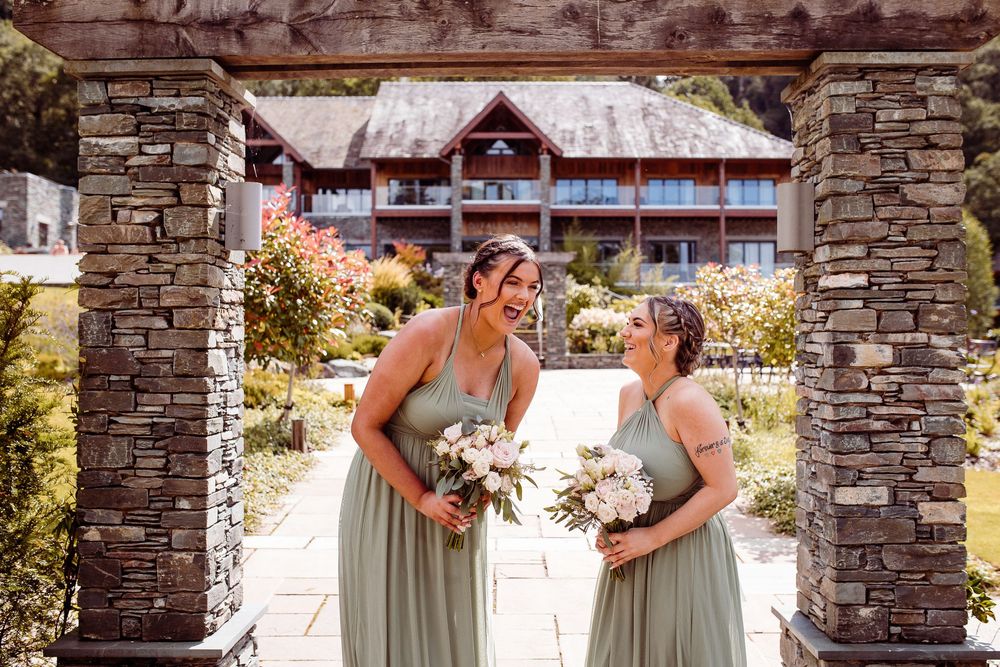 Two bridesmaids in sage green gowns holding bouquets smile at each other under a stone arch at an elegant venue.