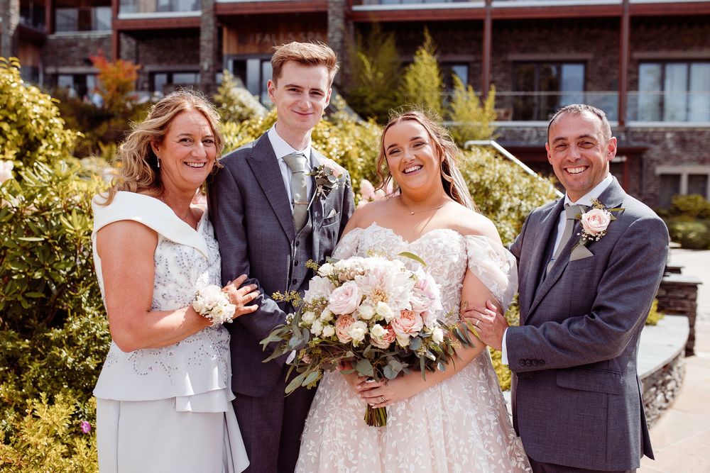 Bride in lace gown holding pink bouquet poses with groom and family outdoors at elegant wedding venue.