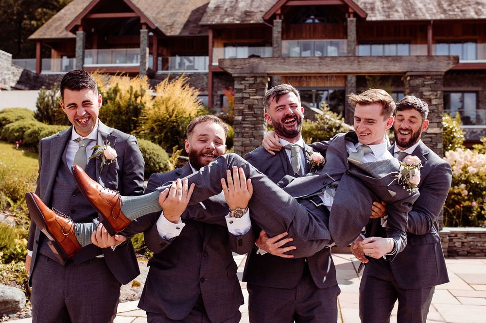 Groomsmen in navy suits laughing while carrying the groom outdoors at a wedding venue.