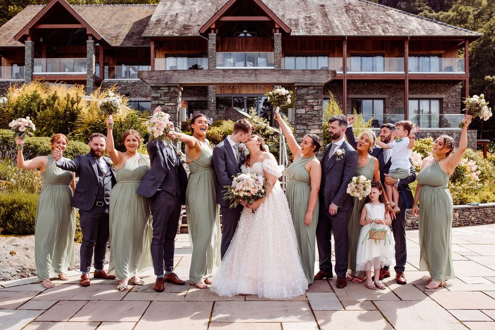 Wedding party celebrating outdoors in sage green bridesmaid dresses and navy suits before a rustic lodge venue.