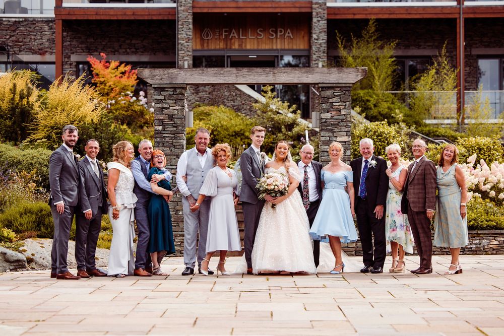 Wedding party group photo outside a modern venue, bride in white gown with groom and family in formal attire.