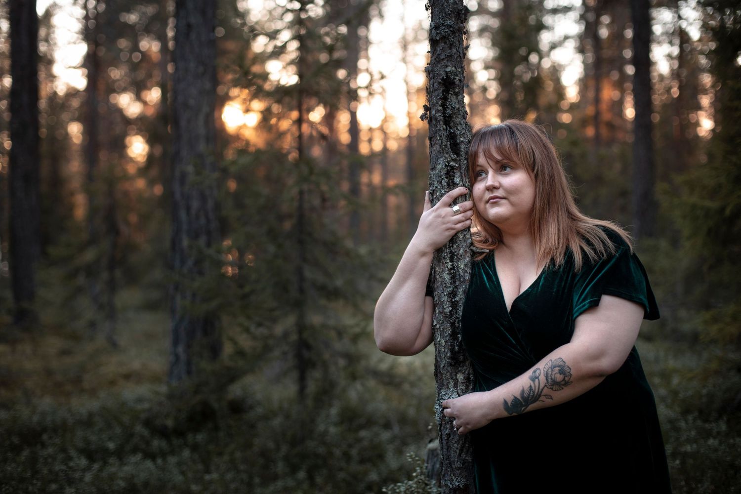 Woman with tattoos leaning against pine tree in forest during golden sunset light in Scandinavian woodland.
