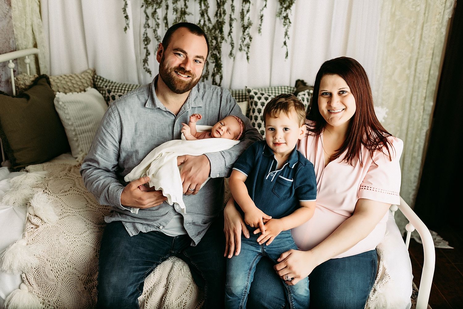 Happy family of four posing for newborn portrait, parents holding baby and toddler son, sitting on cozy bed.