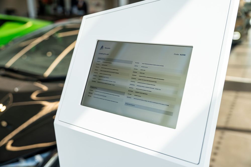 White digital kiosk with touchscreen display in a modern car showroom with luxury vehicles in background.