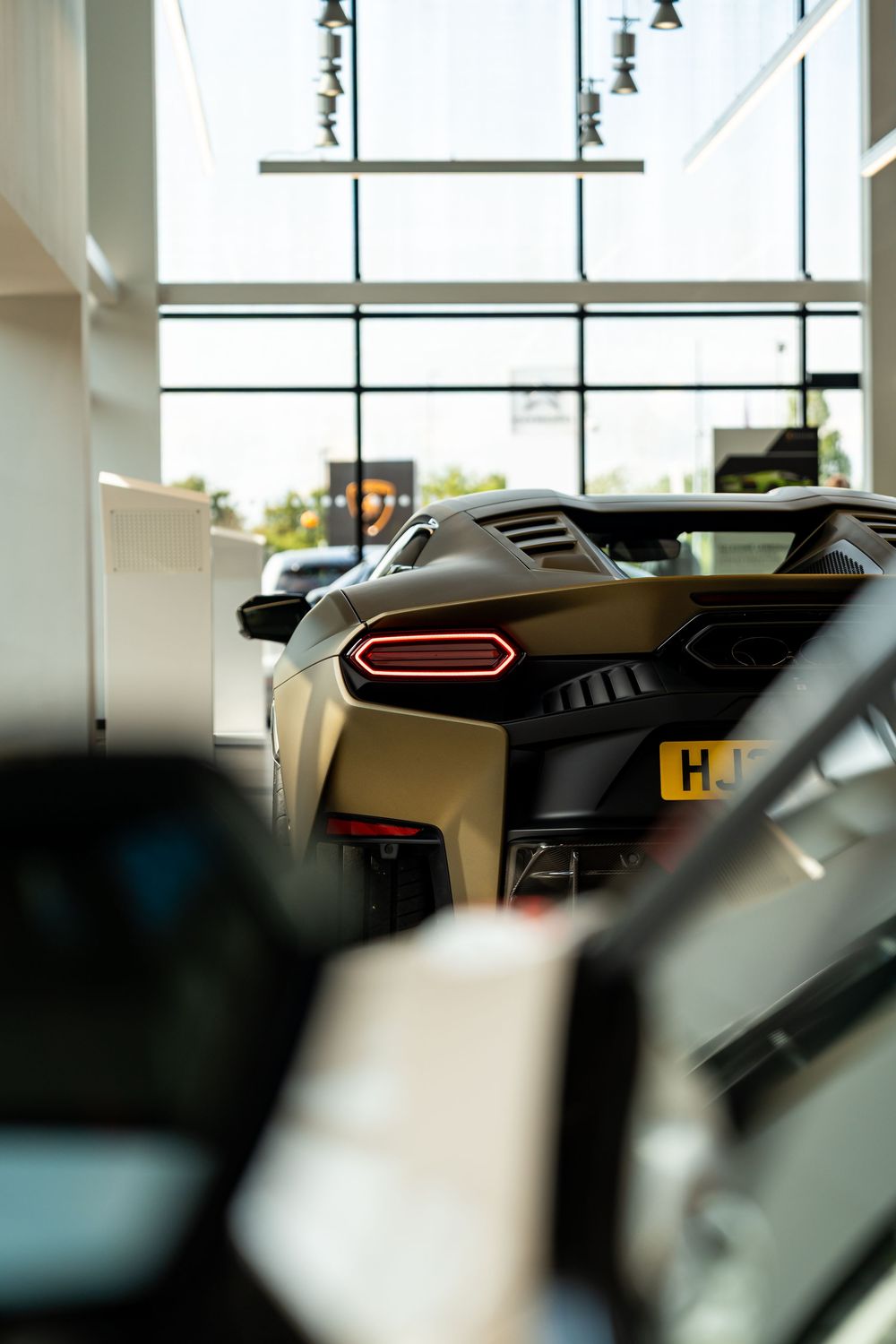 Rear view of a matte gold Lamborghini supercar with red taillights inside a bright showroom.