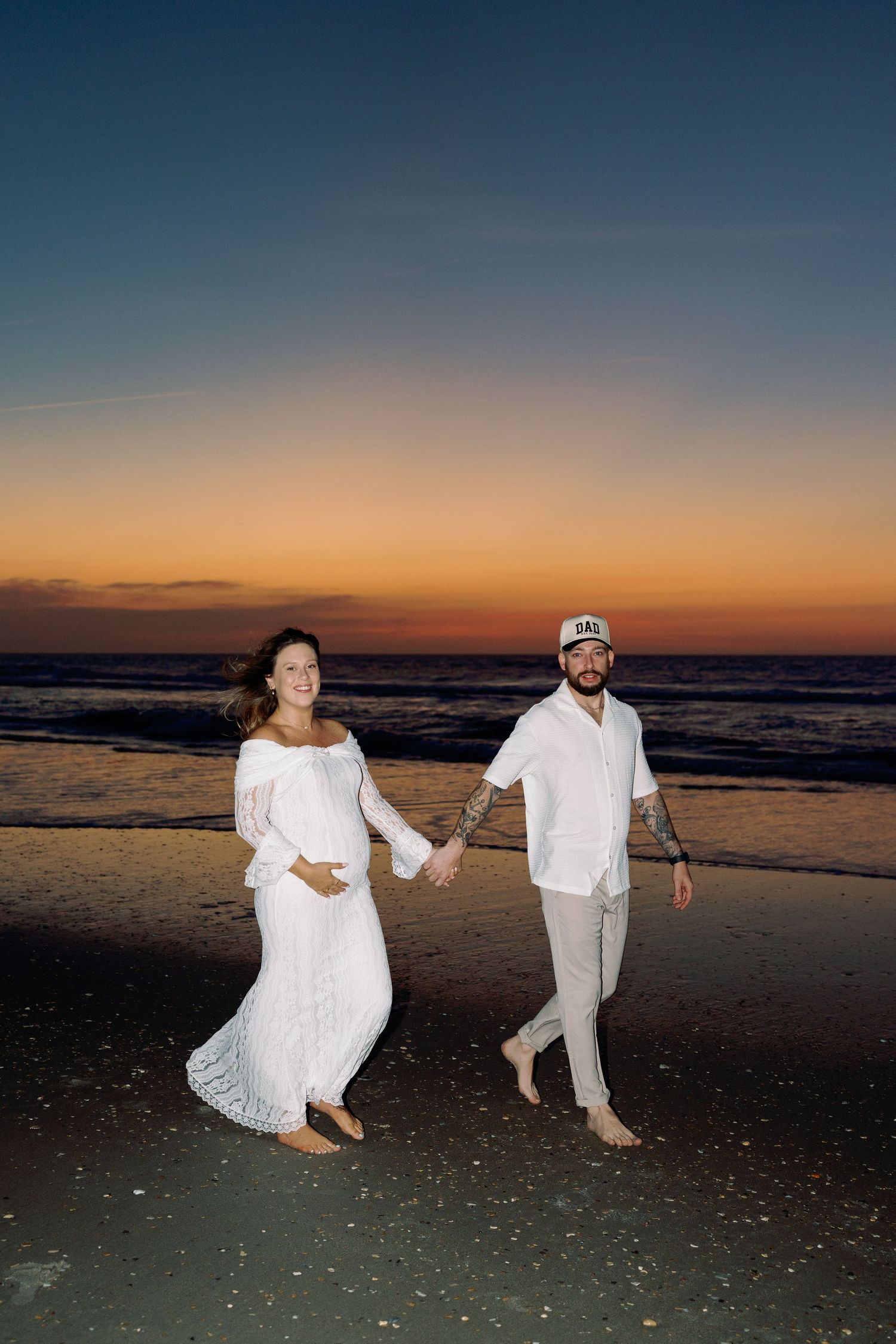 Pregnant couple in white outfits holding hands on beach at sunset, woman in flowing dress, man in cap.