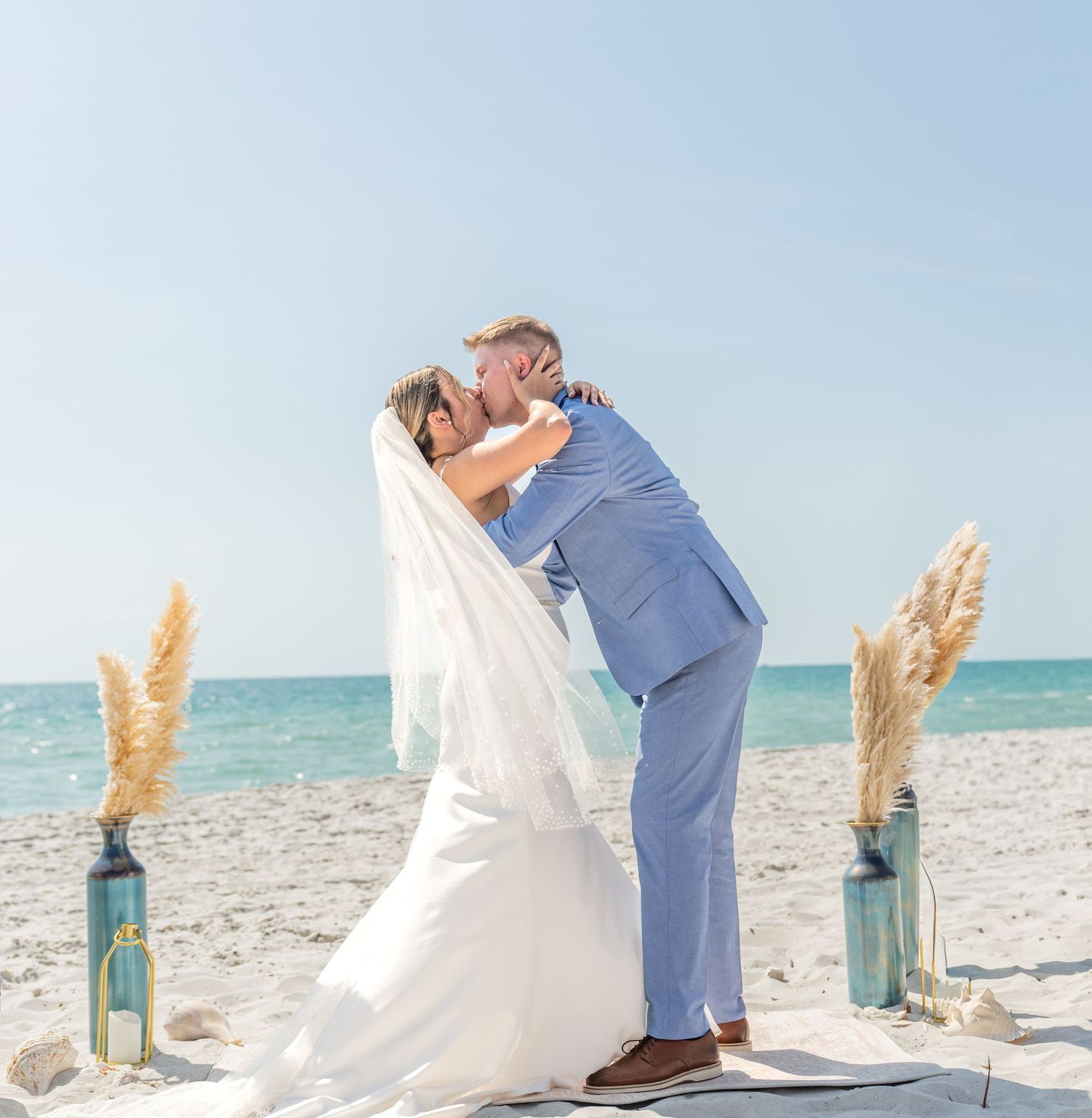 Couple sharing first kiss on sandy beach wedding ceremony with pampas grass decor and ocean backdrop.