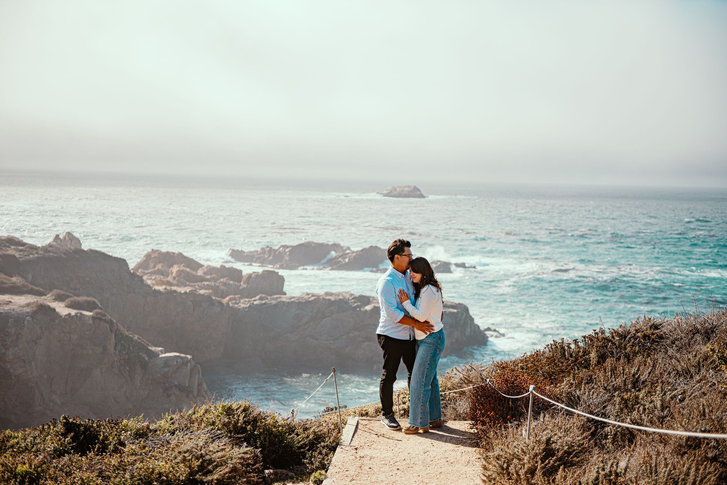 Couple embracing on a coastal cliff path overlooking rocky shores and blue ocean waves on a misty day.