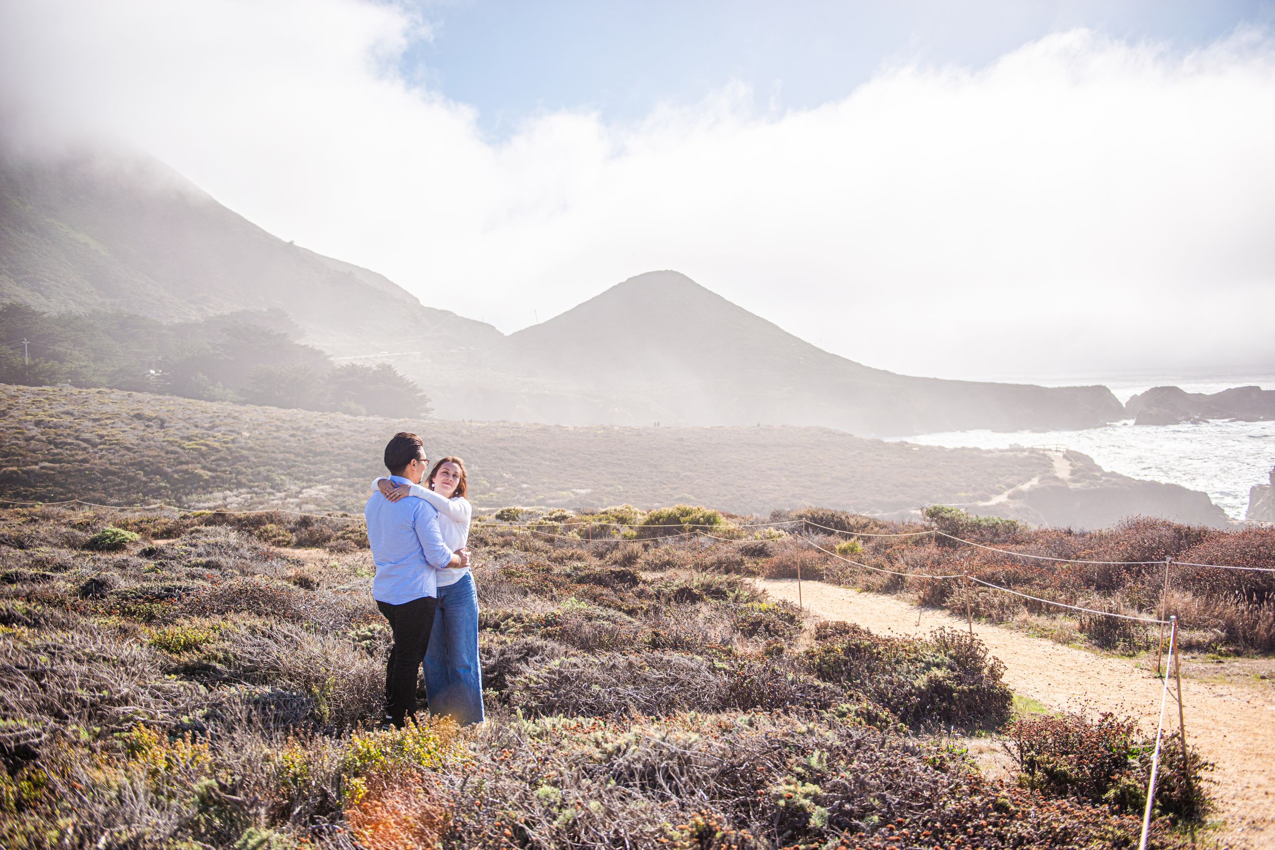 Couple embracing on a scenic coastal trail surrounded by shrubs with misty mountains and ocean in the background.