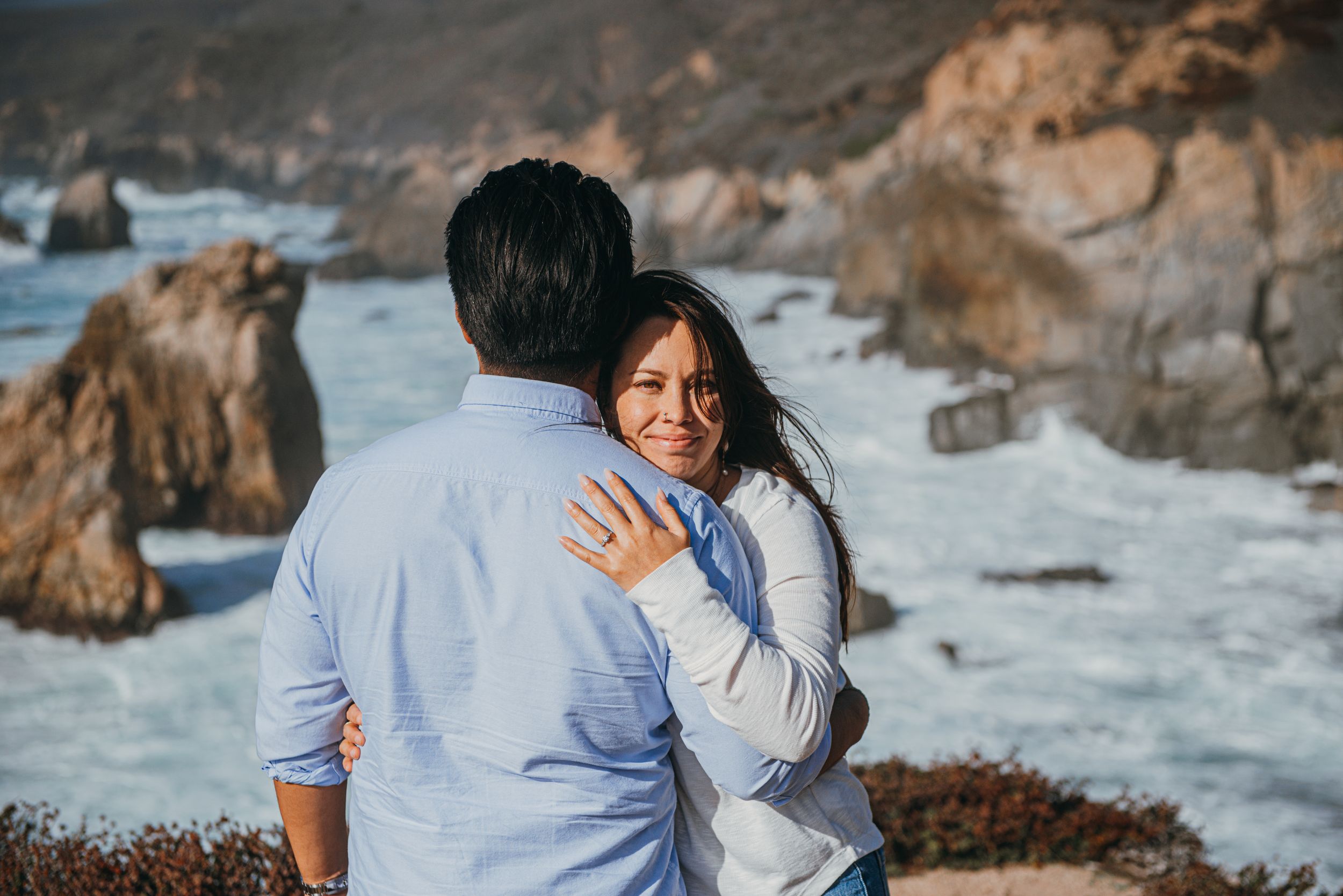 Couple embracing on rocky Big Sur coastline, woman smiling over mans shoulder with ocean waves and cliffs behind them.
