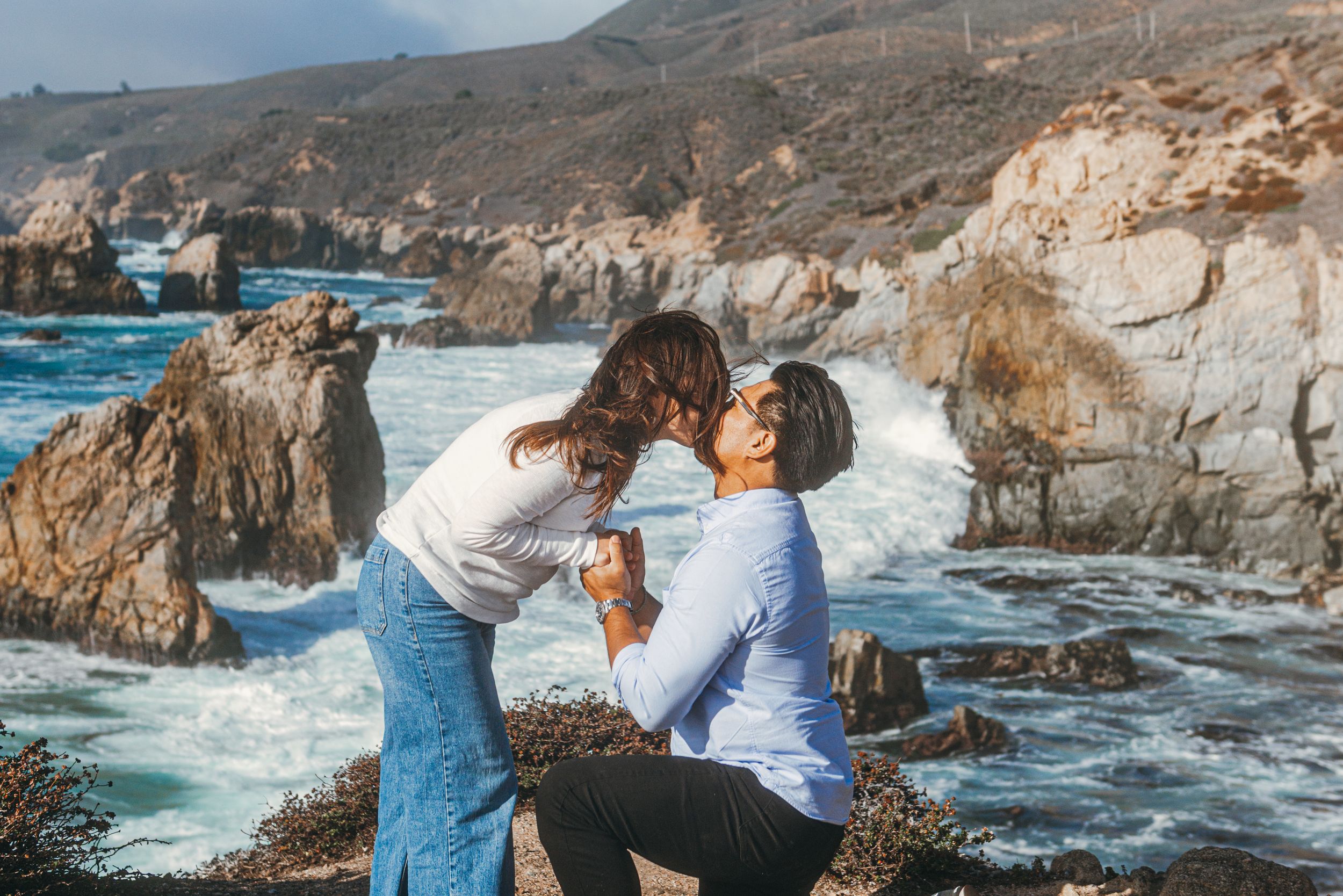 Couple sharing a romantic kiss on rocky coastal cliffs with crashing ocean waves in the background.
