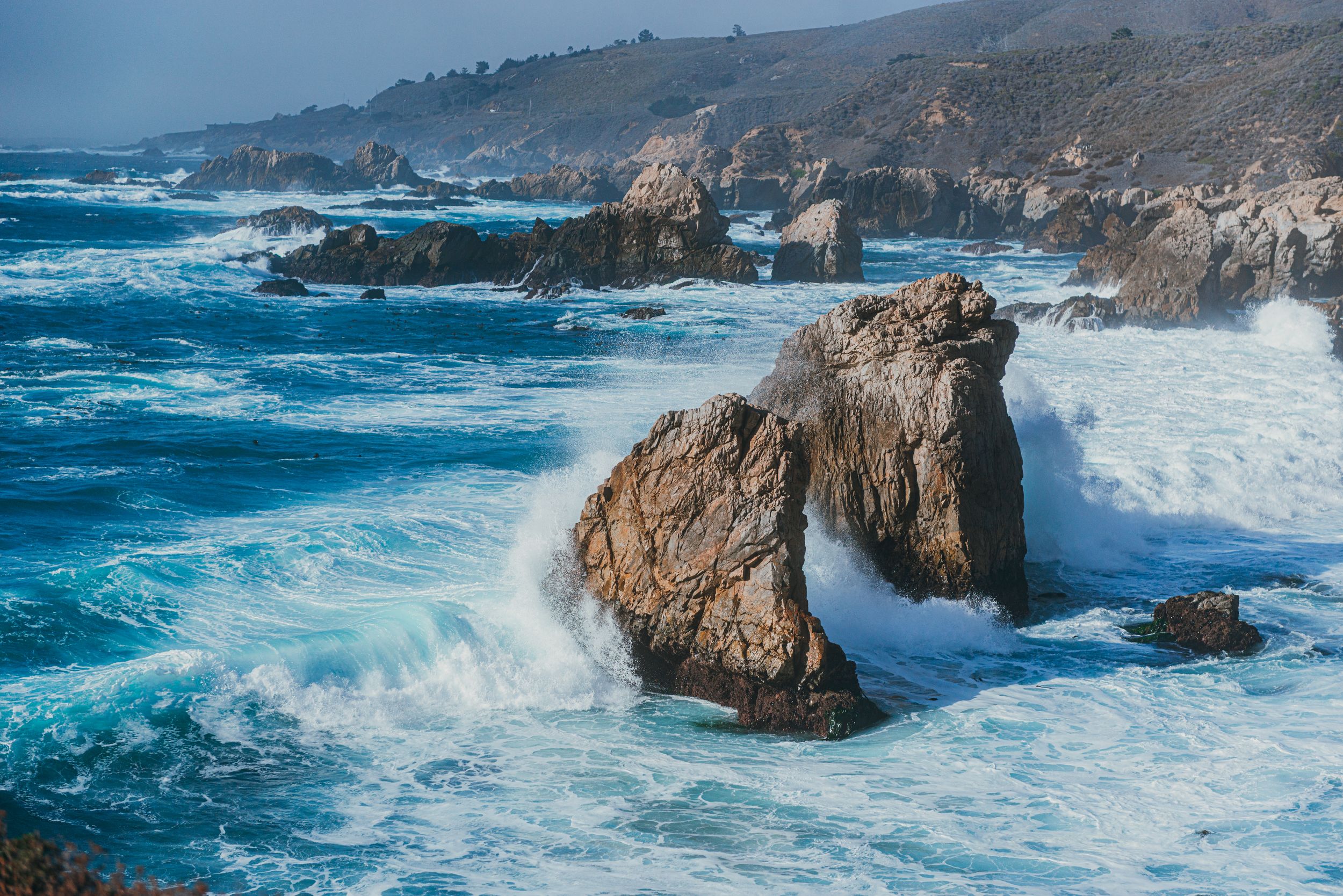 Powerful ocean waves crashing against rocky coastal cliffs with turquoise blue water and misty sea spray.