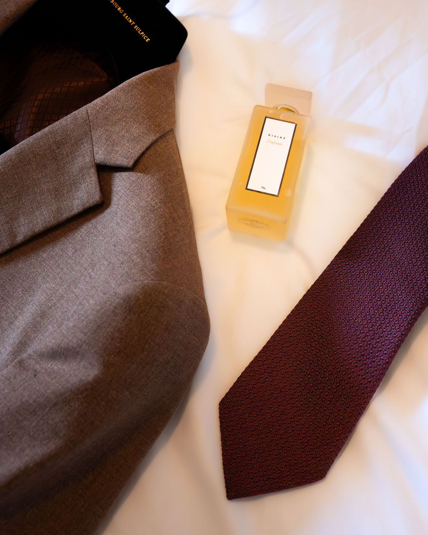 Flat lay of a grey blazer, burgundy tie, and yellow fragrance box on a white surface.
