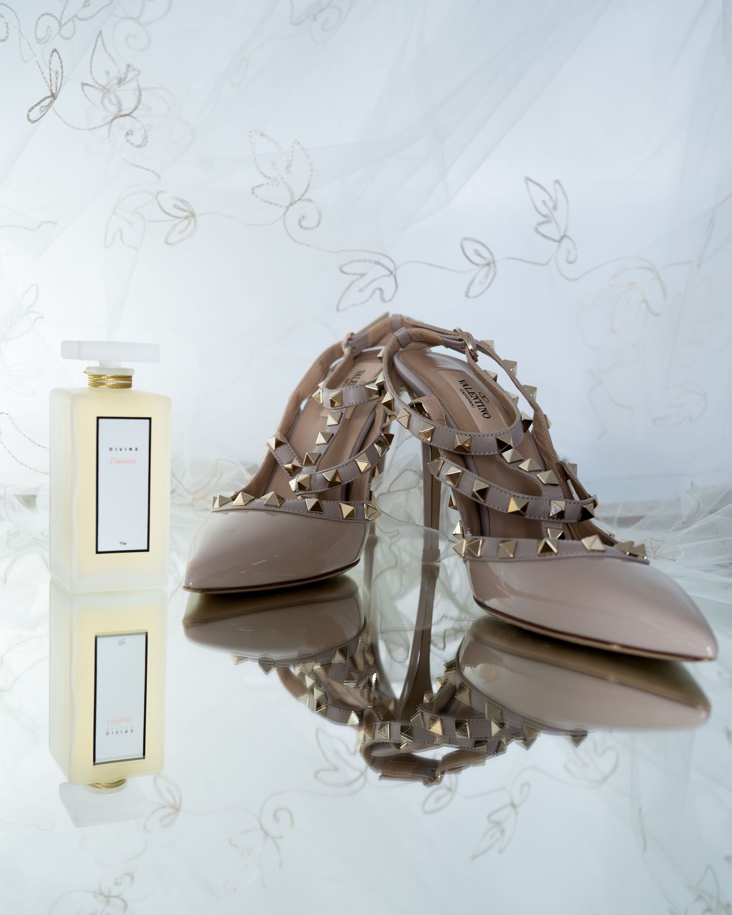 Des escarpins nude Valentino Rockstud se reflètent sur une surface brillante, à côté d'un flacon blanc du parfum Divine. Photo Stph Lecaude