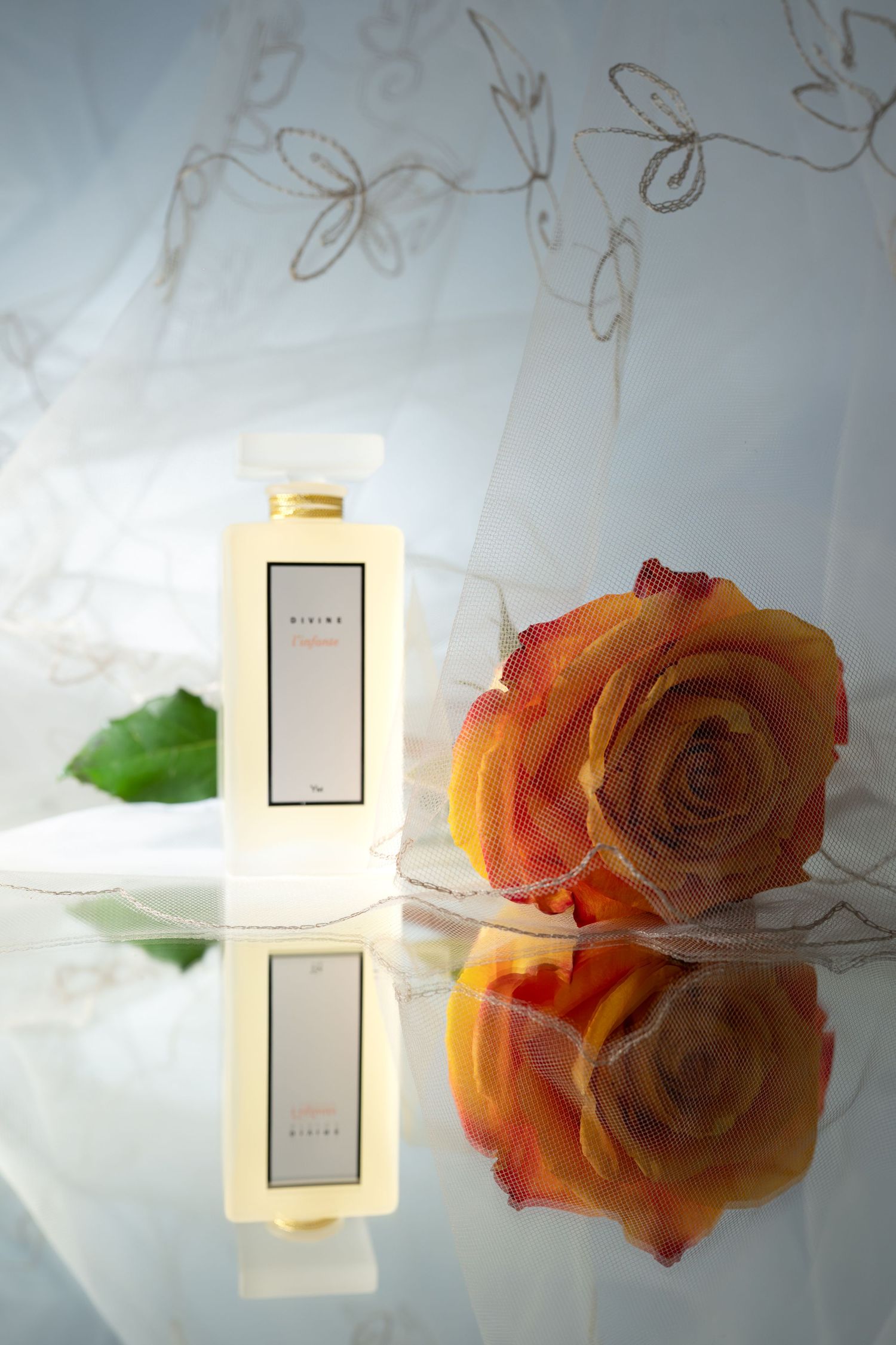 Divine perfume bottle beside an orange rose reflected on a mirrored surface with floral sheer fabric.