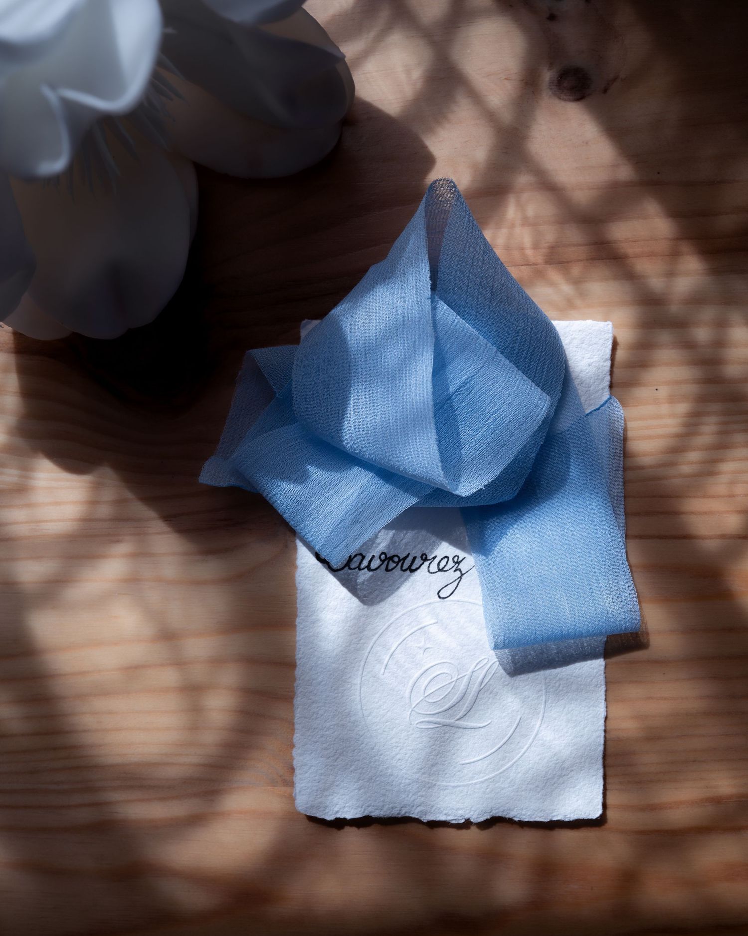 Blue linen ribbon and white embossed card with cursive text on a wooden surface in soft sunlight.