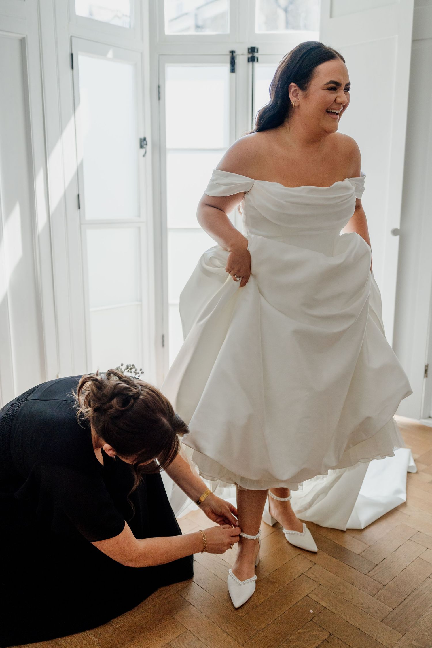Bride in off-shoulder white gown laughing as bridesmaid kneels to fasten her white heels on wedding day.