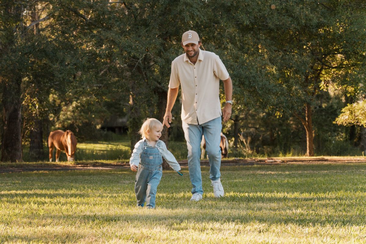 Dad and toddler playing tag on green grass in a sunny farm with trees and a horse in the background during a family photoshoot in Sydney