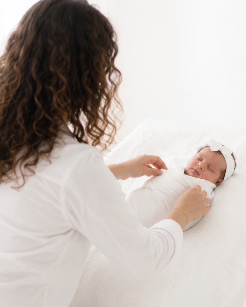 Curly-haired mother in white adjusts her swaddled newborn baby girl wearing a white bow headband.