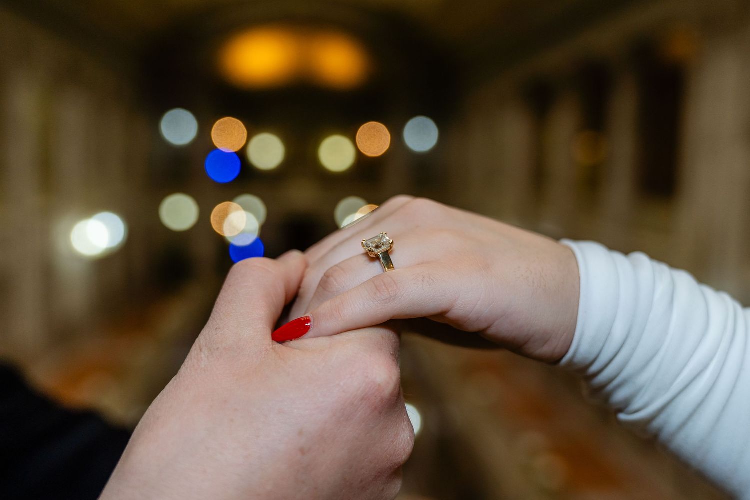 Close-up of two hands holding, woman wearing a gold ring with red nail polish, bokeh lights in background.