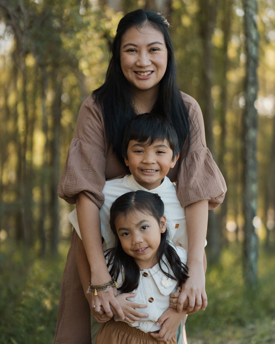 Smiling woman poses outdoors with two young kids in a rouse hill forest setting, all wearing neutral-toned outfits during family photoshoot