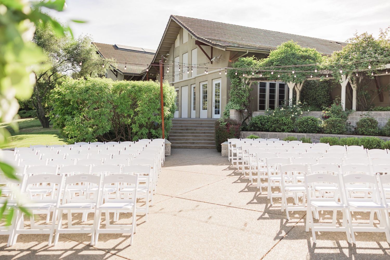 Outdoor wedding ceremony setup with white folding chairs on a sunny patio in front of an elegant venue.