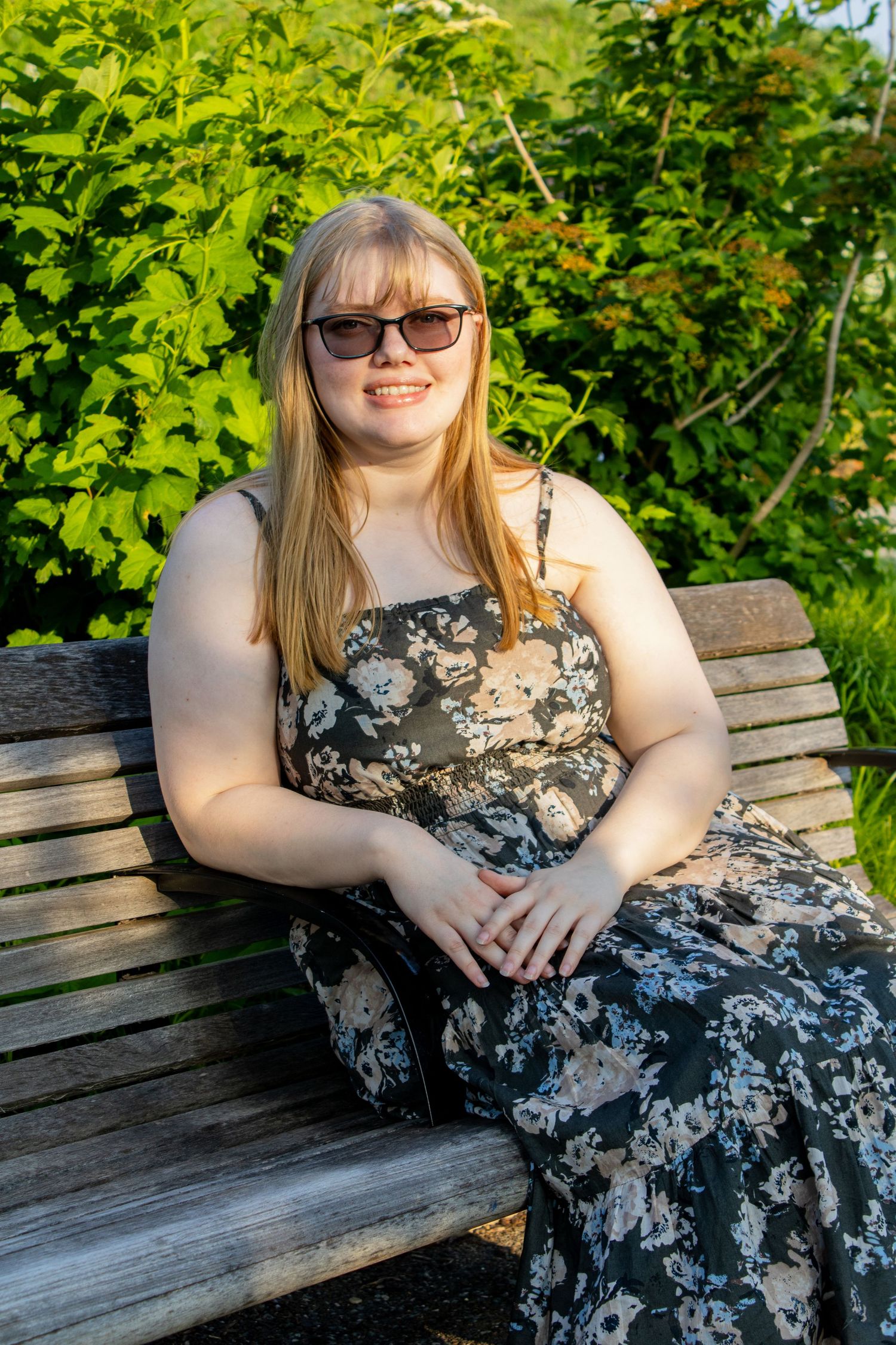 Young blonde woman in floral sundress and sunglasses sitting on wooden bench surrounded by green foliage.
