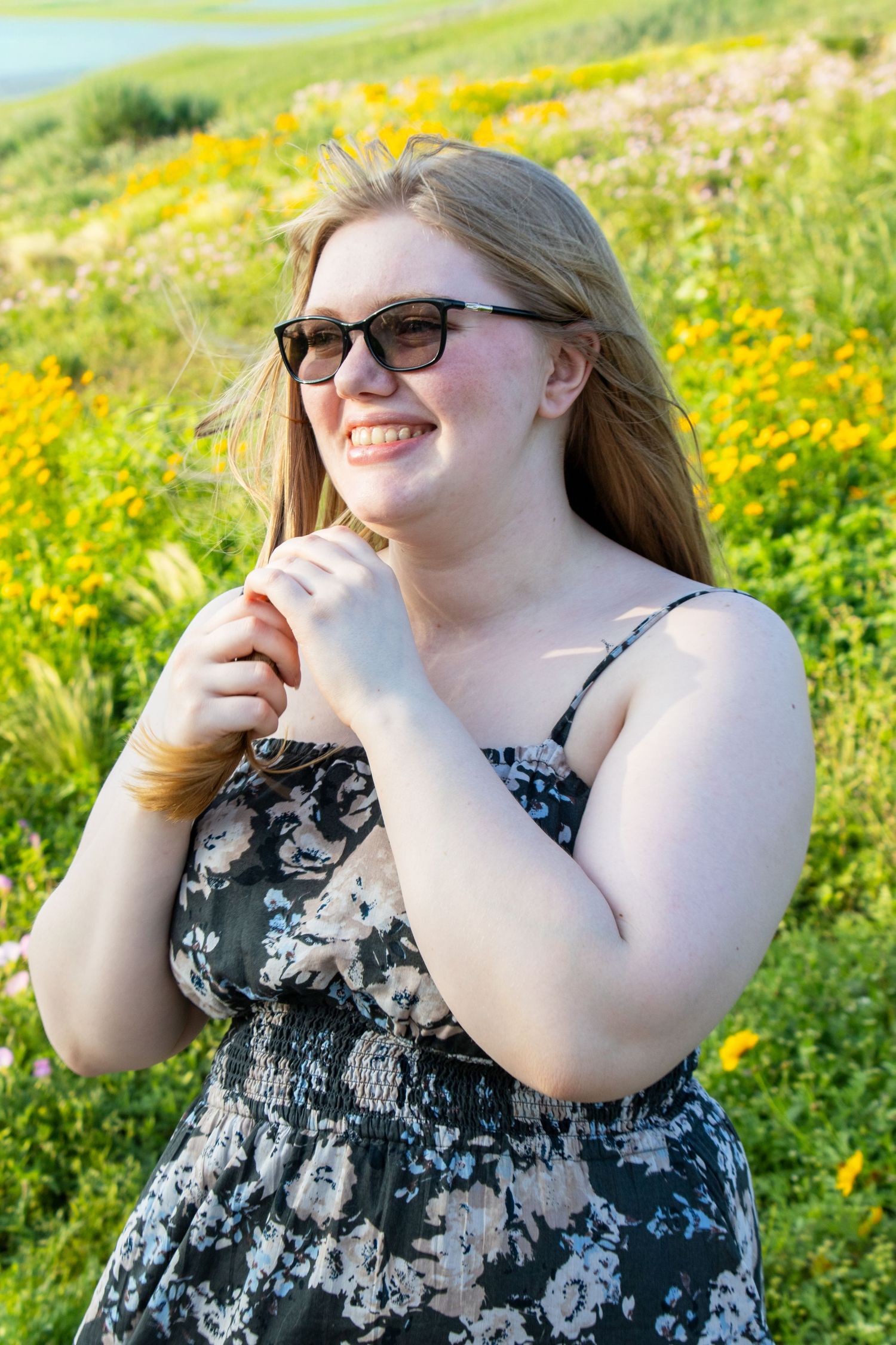 Happy young woman in floral dress and sunglasses standing in a vibrant yellow wildflower meadow.