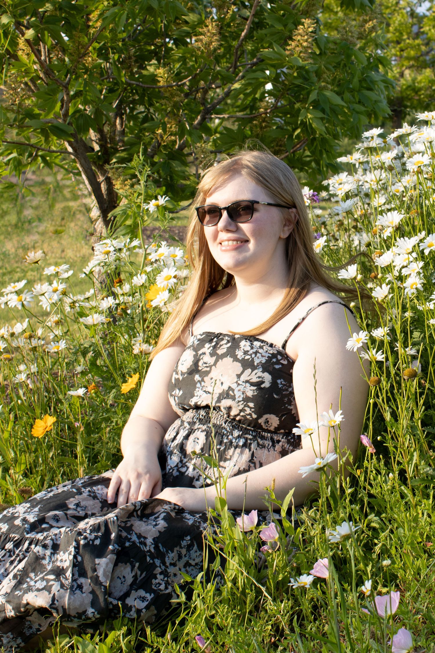 Smiling young blonde woman in floral dress and sunglasses sitting among white and pink wildflowers outdoors.