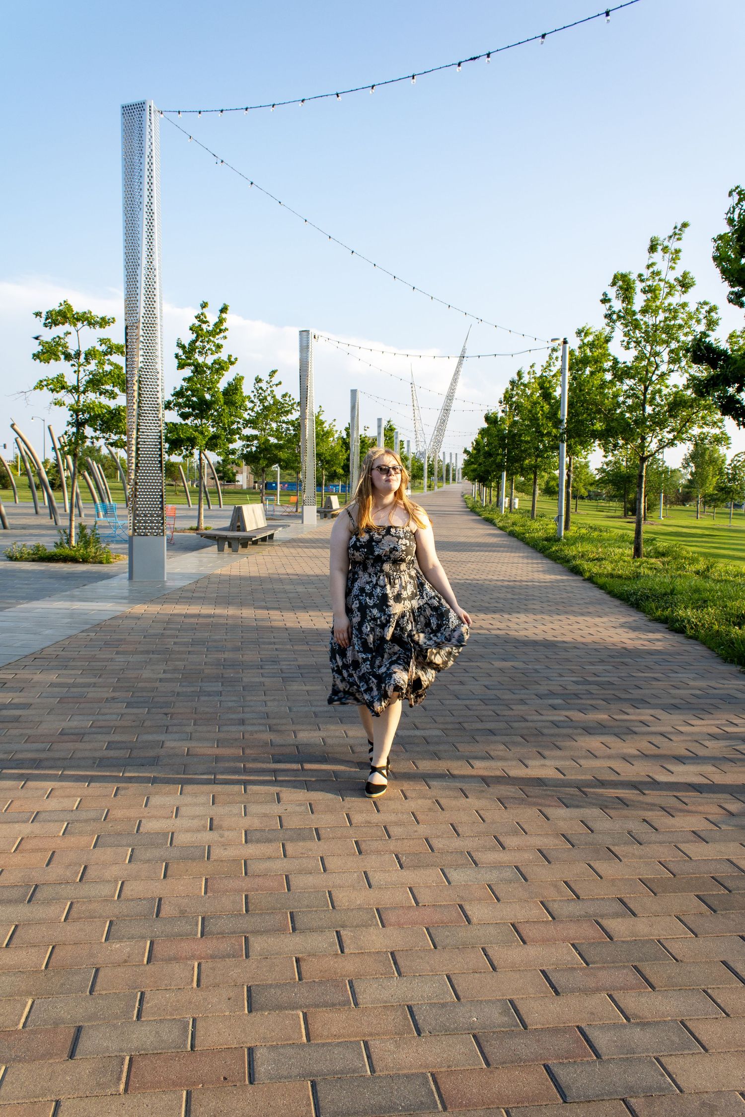Young woman in floral sundress walking along a brick pathway in a sunny park with string lights.