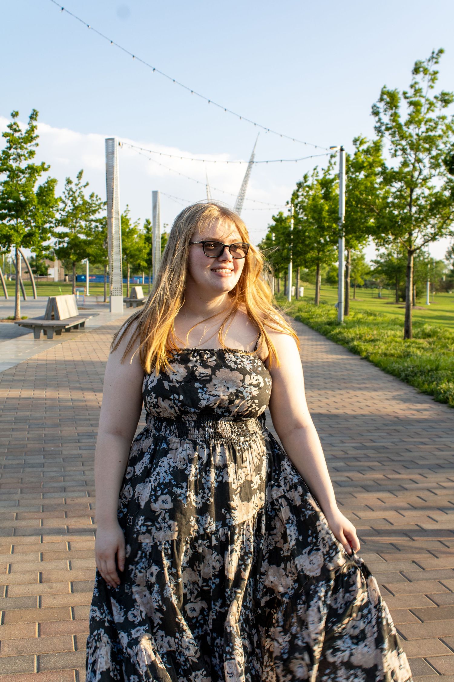 Smiling young woman in floral maxi dress and sunglasses walking on a brick path in a sunny park.