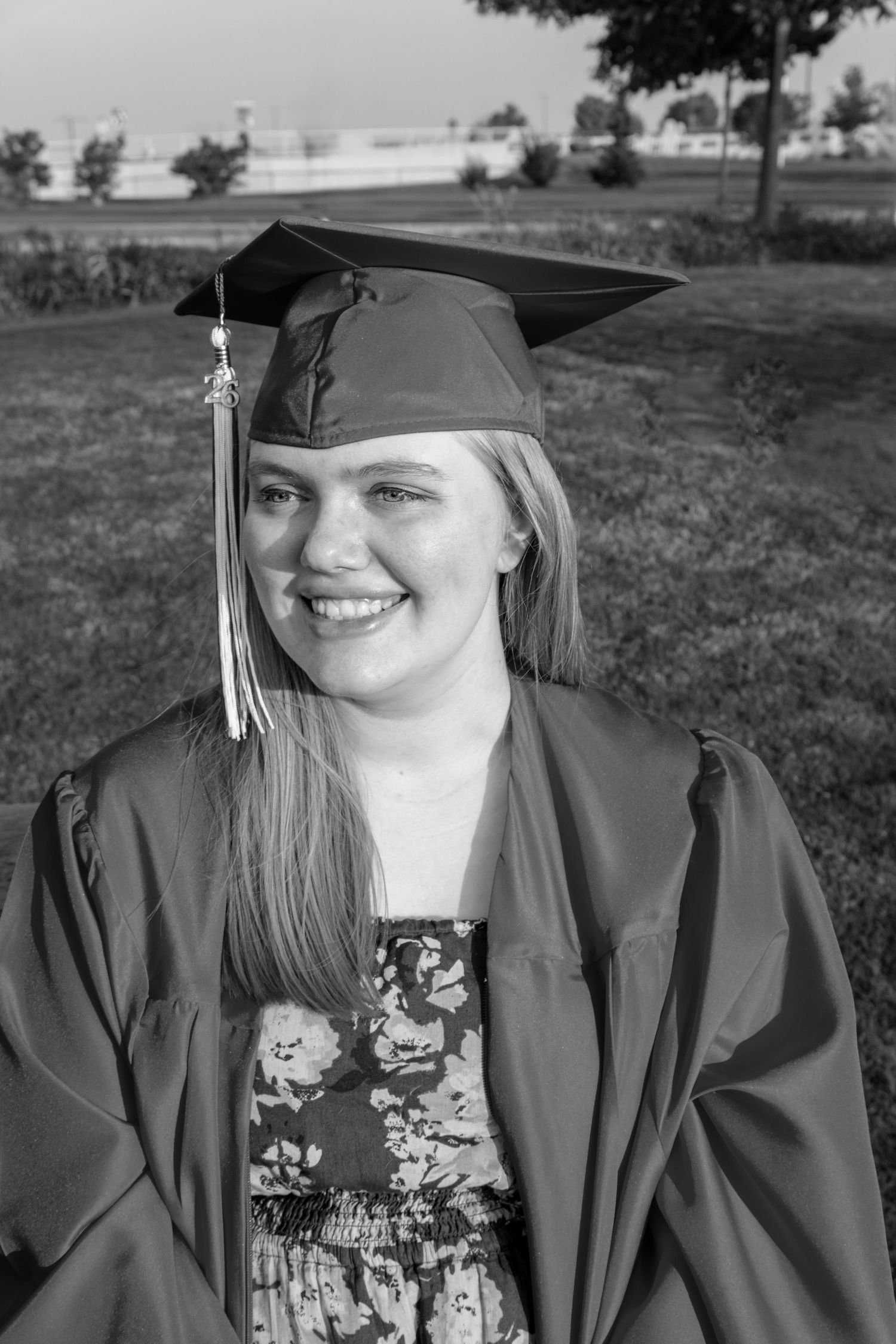 Black and white portrait of smiling young woman in graduation cap and gown outdoors on a sunny day.
