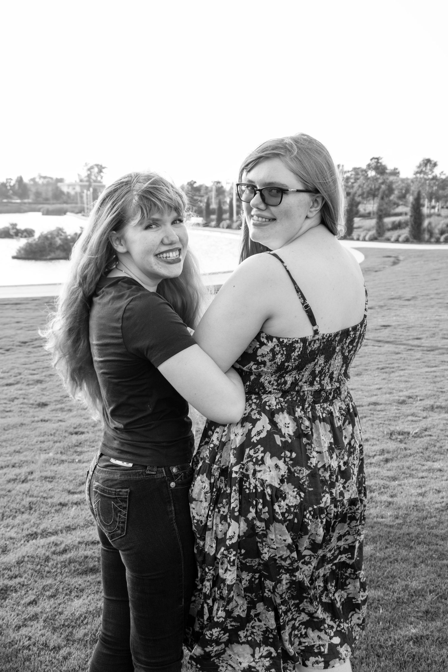 Black and white photo of two smiling young women embracing outdoors near a lake at sunset.