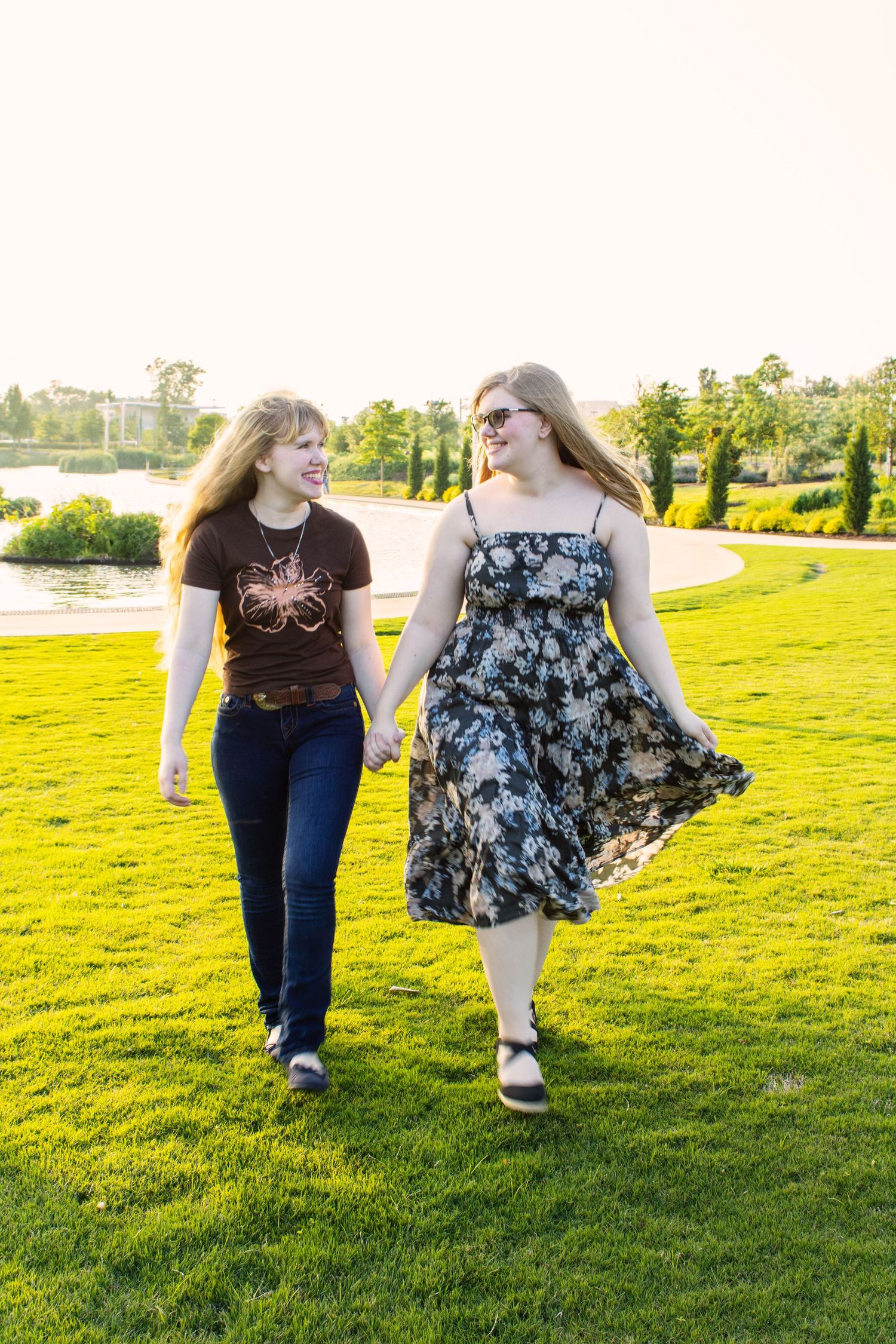 Two young women holding hands and walking together on a sunny green lawn near a scenic pond.